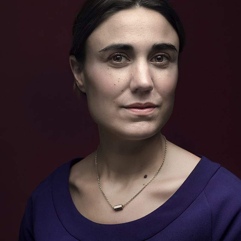 Intieme narratieven 4 Fine art zakelijk portret van een vrouw met donker haar in een losse knot en donkerblauwe blouse, kijkend in de camera tegen een donkere achtergrond.