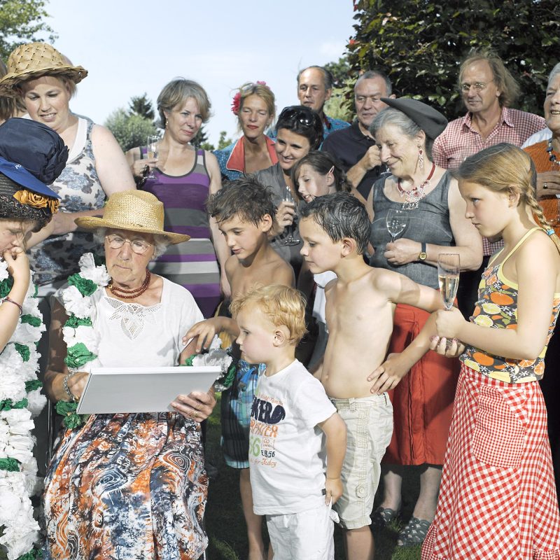 Zo'n achttien personen van kind tot oudere staan samen in een tuin; de aandacht gaat naar een oudere vrouw met strohoed die een wit voorwerp vasthoudt.