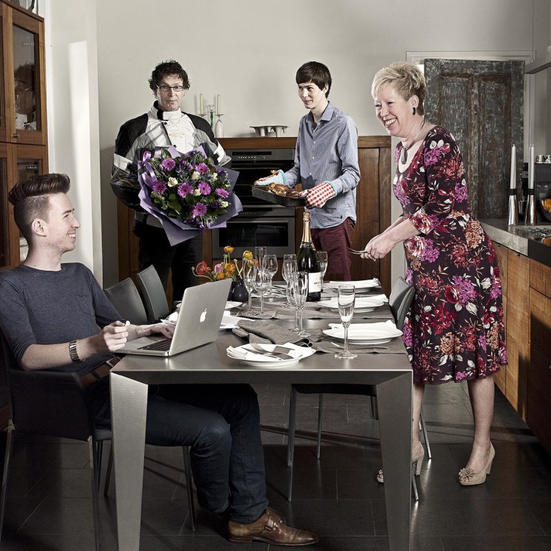 Vier personen van verschillende leeftijden in een moderne keuken rond een eettafel; één werkt aan een laptop, één houdt een boeket bloemen, één een schaal eten.