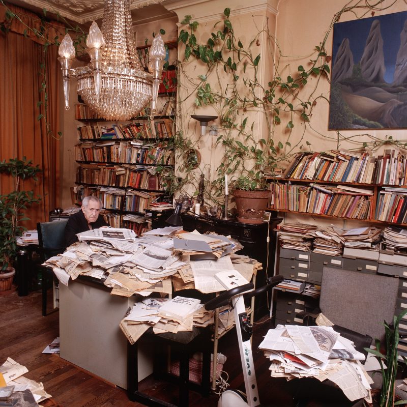 Intieme narratieven 3 Frits Frenkel.Oudere schrijver met grijs haar en bril in donkere trui aan een bureau vol stapels papier, in een werkkamer met boekenkasten en kroonluchter.