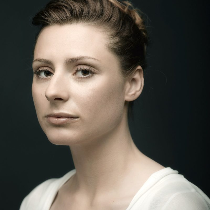 Intieme narratieven 2 Fine art portret van een jonge vrouw met donker haar in een losse knot, in witte blouse tegen een zwarte achtergrond.