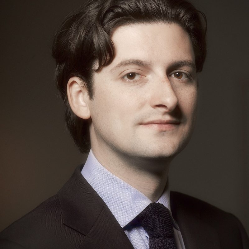 Intieme narratieven 1 Portret van Jan Six, een man in donker pak met lichtblauw overhemd en das, kijkend in de camera tegen een donkere egale achtergrond.
