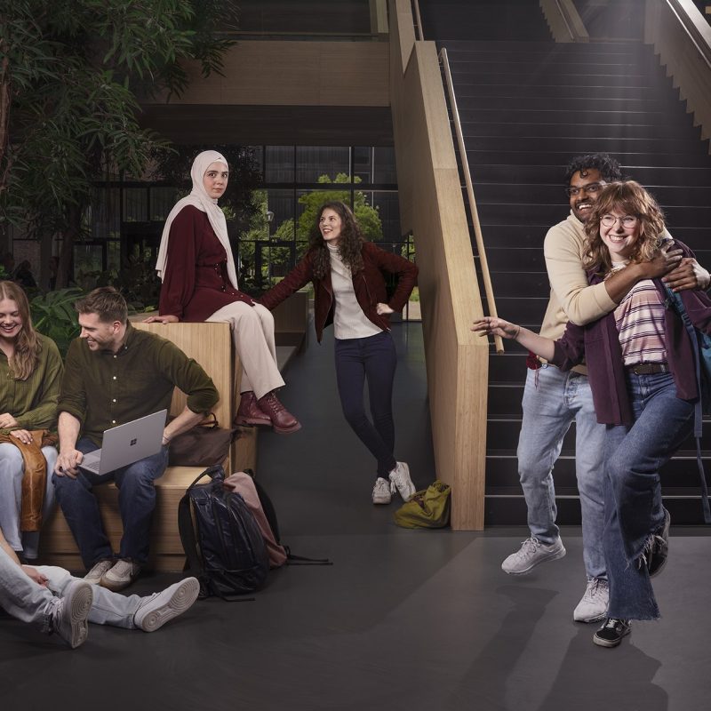 Tableau vivant 7 Acht studenten in een moderne campusruimte met trap en planten; rechts tillen twee elkaar speels op, links zit een groep bij elkaar op bank en grond.