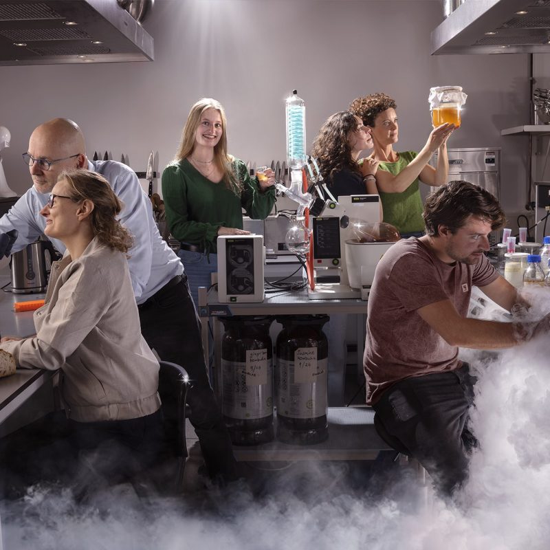 Tableau vivant 8 Zes studenten en docenten heffen glazen vloeistof in een keuken-laboratorium vol damp en wetenschappelijke apparatuur, op de campus van Hogeschool Leiden.