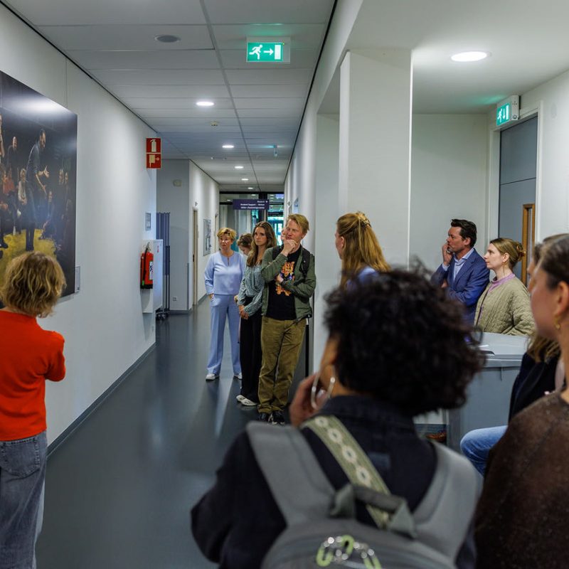 Een groep bezoekers staat in een gangpad van een expositieruimte en kijkt naar een groot kleurrijk kunstwerk met gele accenten aan de muur.