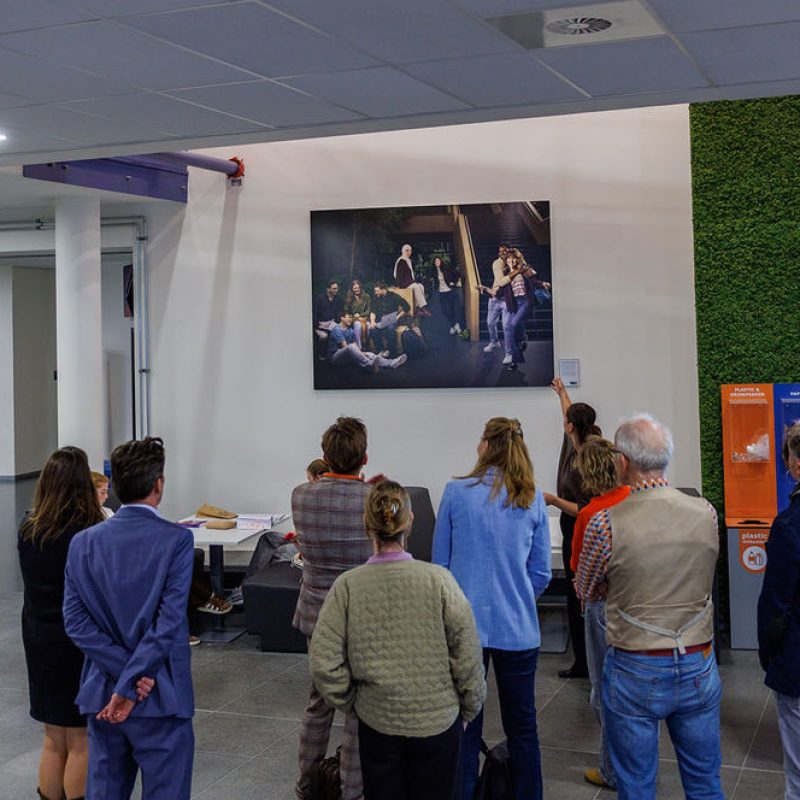 Zo'n vijftien exposanten bij een expositie kijken naar een grote zwart-wit foto aan de muur, naast kleurrijke posters tegen een groene wand.
