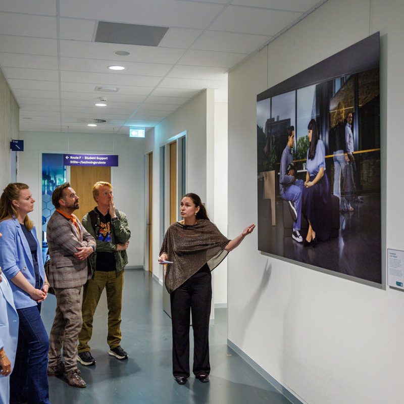 Een groep van acht bezoekers luistert in een gang naar een vrouw die uitleg geeft bij een grote kleurrijke foto aan de muur.